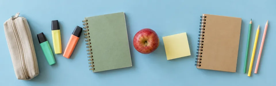 A pencil case, highlighters, spiral notebooks, an apple and a sticky-note pad on a sky-blue table.