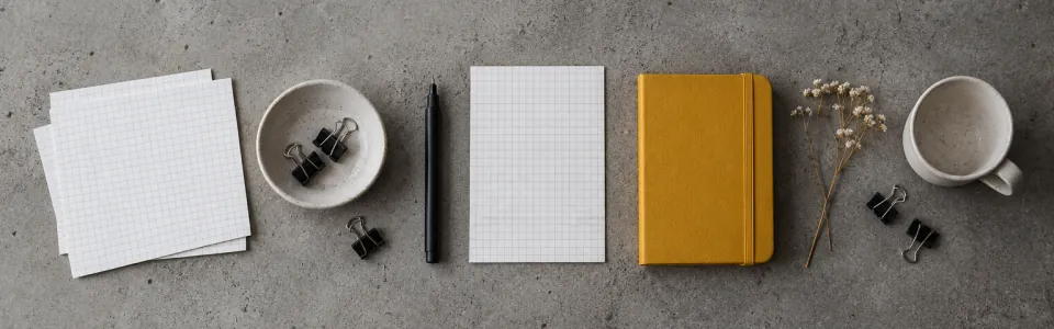 Grid paper sheets, a black pen, binder clips, a mustard notebook and a cup on grey concrete.