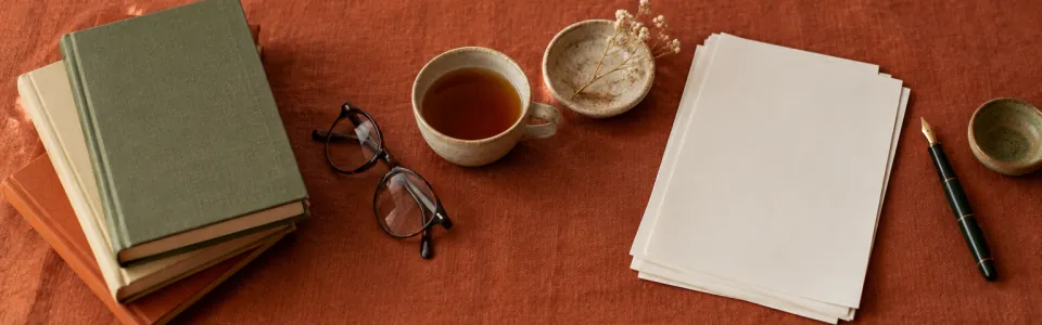 A stack of books, glasses, a cup of tea, loose pages and a fountain pen on terracotta cloth.